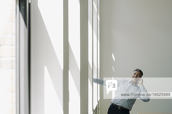 Happy businessman talking on mobile phone while hanging against wall in office