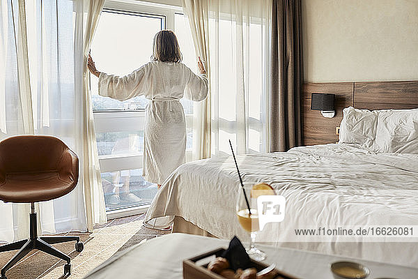 Ältere Frau im Ruhestand öffnet den Vorhang und blickt durch das Fenster eines luxuriösen Hotelzimmers