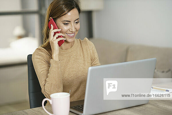 Lächelnde Geschäftsfrau  die am Telefon spricht  während sie zu Hause am Laptop arbeitet