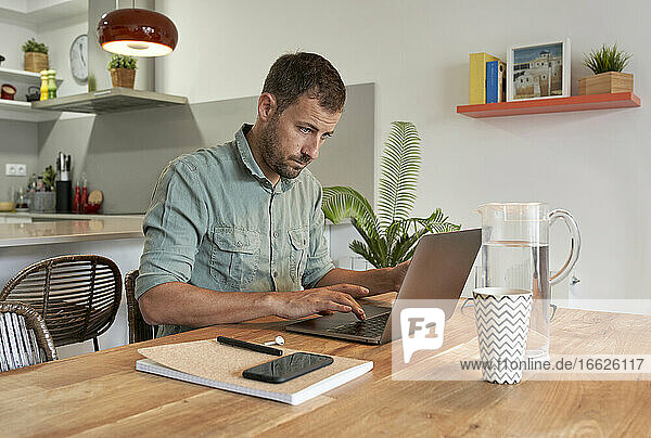 Gutaussehender männlicher Freiberufler mit Laptop am Esstisch bei der Arbeit von zu Hause aus
