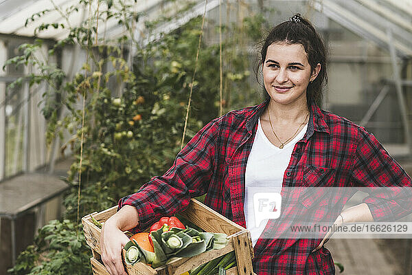 Junge Frau  die eine Kiste mit Gemüse trägt und gegen ein Gewächshaus blickt