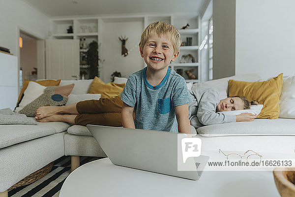 Lächelnder Junge mit Laptop und schlafender Mutter im Hintergrund