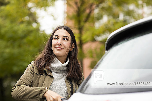 Schöne Frau lädt gemietetes Elektroauto auf  während sie lächelt und wegschaut