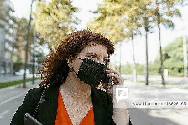 Frau mit Gesichtsmaske  die auf einer Straße in der Stadt steht und mit einem Mobiltelefon spricht