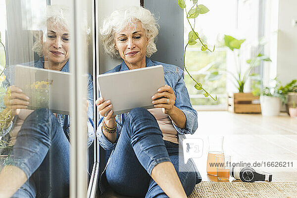 Reflektierendes Bild einer reifen Frau  die zu Hause sitzend an einem digitalen Tablet arbeitet