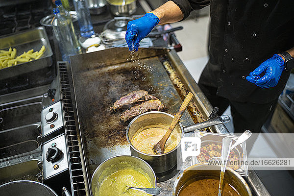 Mann bestreut Fleisch mit Salz  während er am Gasherd in einer Großküche steht