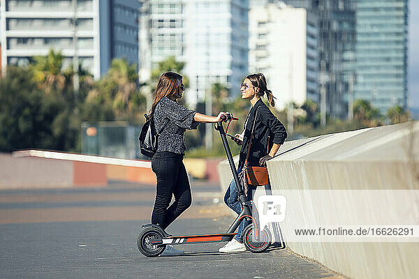 Woman talking with friend while standing with push scooter on road in city