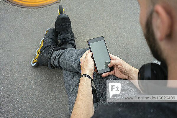 Nahaufnahme eines jungen Mannes  der Inline-Skates trägt und sein Smartphone benutzt  während er auf der Straße sitzt