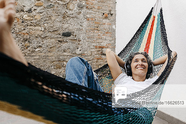 Smiling woman listening music through headphones while relaxing on hammock against wall at back yard