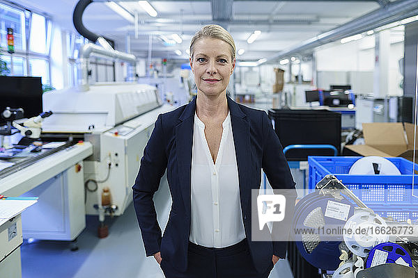 Selbstbewusst lächelnde Geschäftsfrau  die mit den Händen in den Taschen in einer beleuchteten Fabrik steht