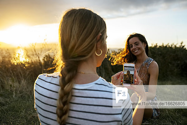 Young woman photographing female friend through smart phone during sunset