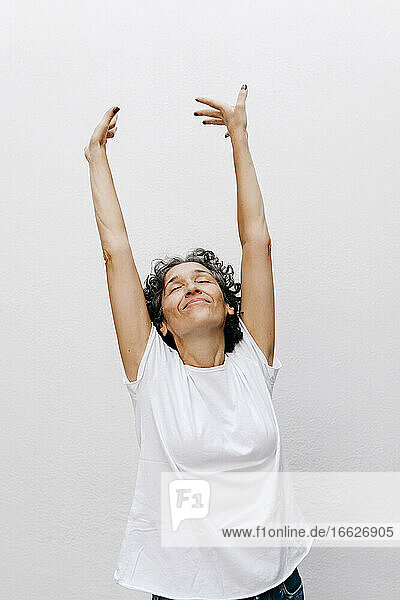 Ältere Frau steht mit geschlossenen Augen und erhobenen Armen vor einer weißen Wand
