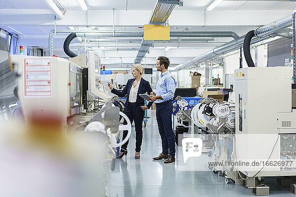 Reife Geschäftsfrau  die mit einem männlichen Ingenieur in einer beleuchteten Fabrik über Maschinen diskutiert