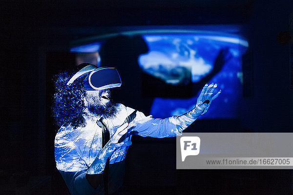 Verspielter Mann mit Virtual-Reality-Brille  der während einer Pandemie zu Hause im Dunkeln steht