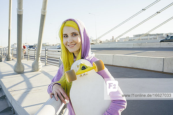Female hipster smiling while holding skateboard standing on bridge