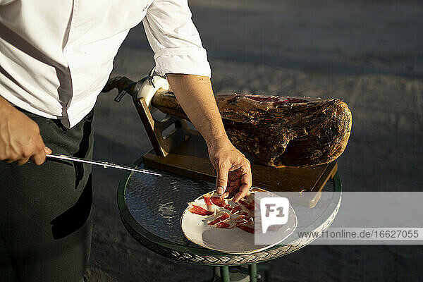 Male chef cutting slices of ham on table while standing outdoors