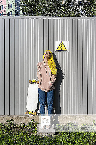Contemplating young woman with hands in pockets standing against metal wall on sunny day