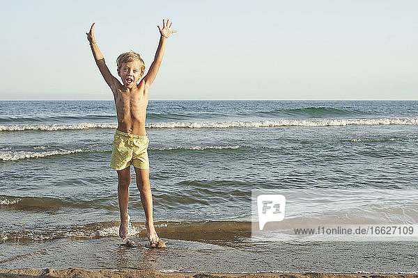 Fröhlicher Junge mit erhobener Hand am Meer stehend am Strand