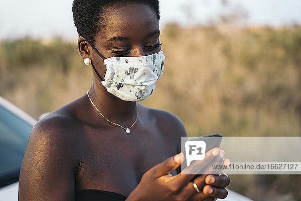 Young woman with face mask using mobile phone while standing at filed