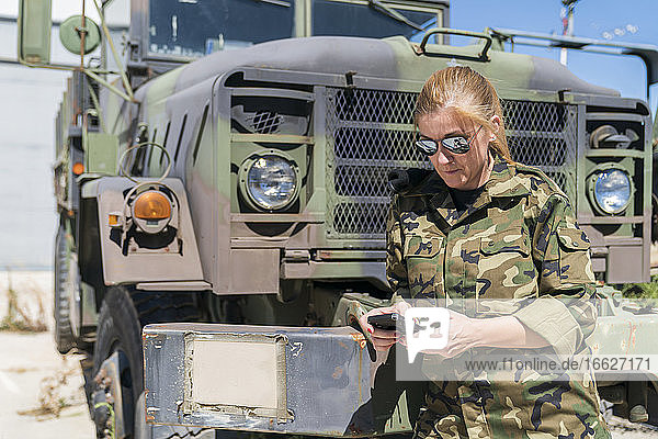Reife blonde weibliche Armee Soldat Text-Messaging durch Smartphone  während stehend gegen LKW an der Basis auf sonnigen Tag