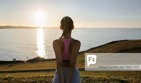 Junge Frau schaut auf das Meer  während sie auf einem Hügel gegen den Himmel bei Sonnenuntergang trainiert