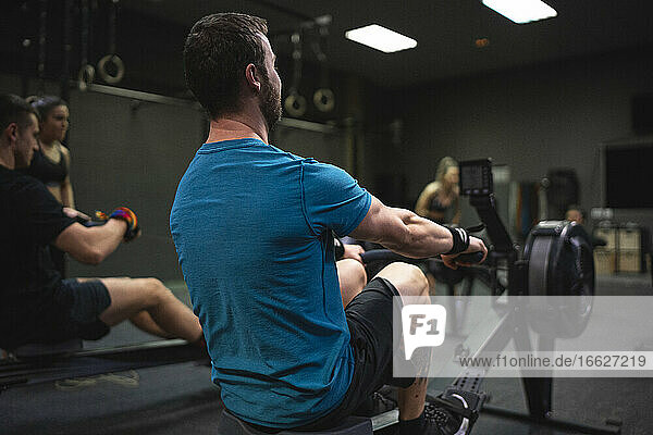 Mann benutzt Rudergerät mit Menschen  die im Hintergrund im Fitnessstudio trainieren