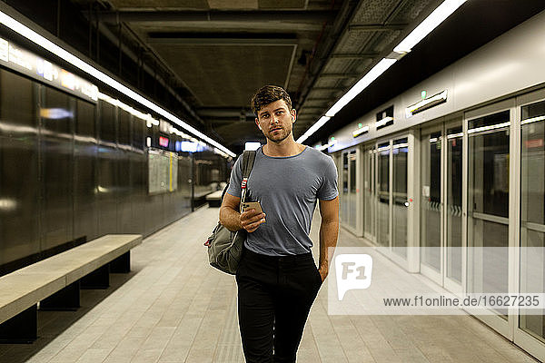 Handsome young man holding smart phone while walking with hand in pocket at illuminated subway platform