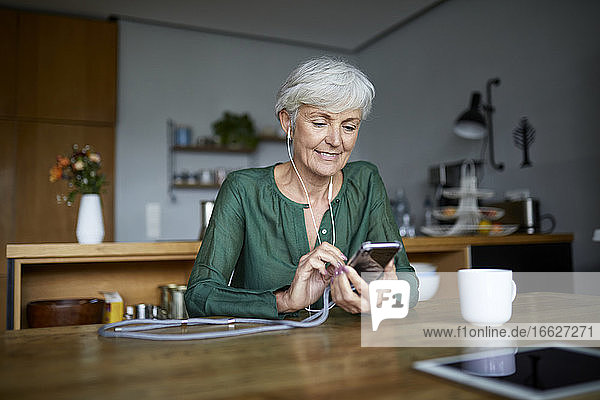 Ältere Frau schreibt Textnachrichten und hört Musik auf ihrem Smartphone  während sie zu Hause sitzt
