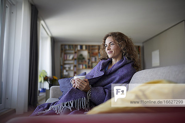 Lächelnde Frau mit Kopftuch  die zu Hause auf dem Sofa sitzt und Kaffee trinkt