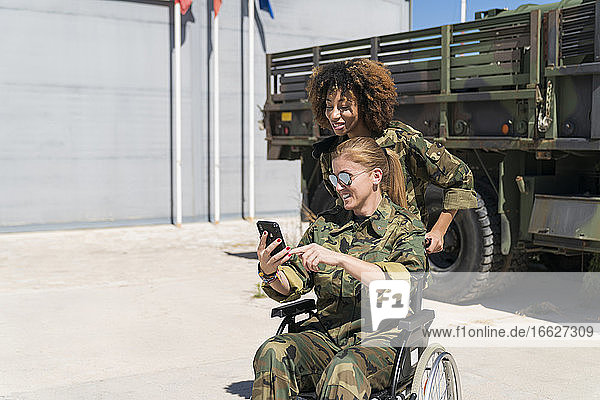 Lächelnde behinderte Soldatin  die ein Selfie mit einem jungen Kollegen vor einem Lastwagen auf einem Armeestützpunkt an einem sonnigen Tag macht