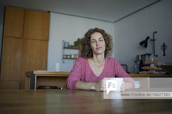 Mittlere erwachsene Frau trinkt Kaffee  während sie zu Hause am Tisch sitzt
