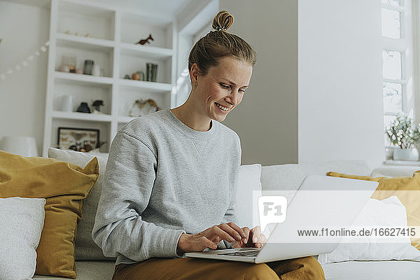 Lächelnde Frau  die einen Laptop benutzt  während sie zu Hause auf dem Sofa sitzt