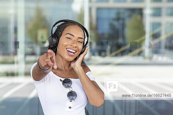 Lächelnde Frau mit Kopfhörer gestikuliert  während sie gegen eine Glaswand in der Stadt steht