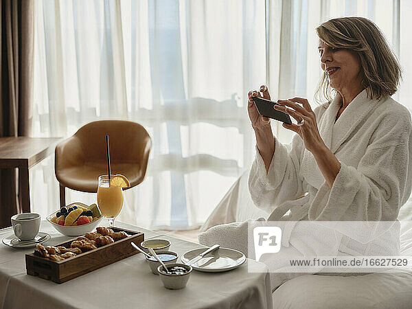 Lächelnde blonde ältere Frau  die das Frühstück mit ihrem Smartphone fotografiert  während sie auf dem Bett im Hotelzimmer sitzt