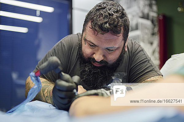 Close-up of bearded artist tattooing on customer's hand in studio