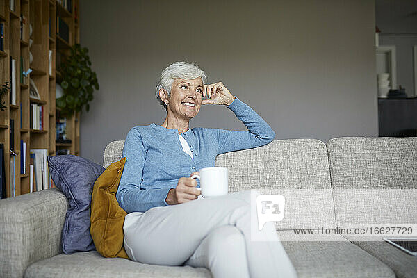 Nachdenkliche ältere Frau  die zu Hause auf dem Sofa sitzt und eine Kaffeetasse hält