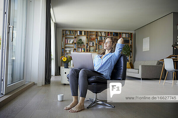 Lächelnde Frau mit Händen hinter dem Kopf  die einen Laptop benutzt  während sie auf einem Stuhl zu Hause sitzt