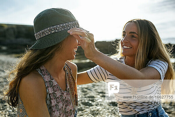 Lächelnde junge Frau  die den Hut ihrer Schwester zurechtrückt  während sie ihr Wochenende am Strand genießt