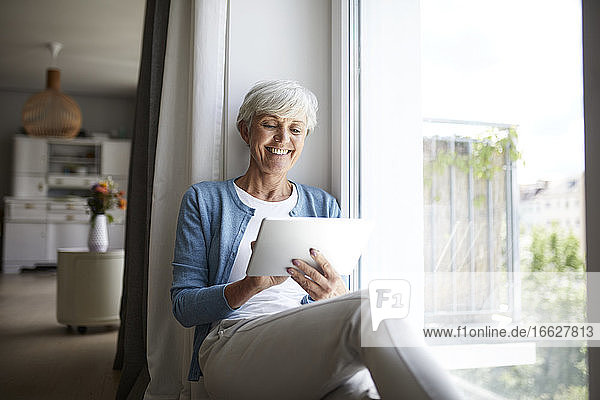 Lächelnde ältere Frau  die ein digitales Tablet benutzt  während sie zu Hause am Fenster sitzt