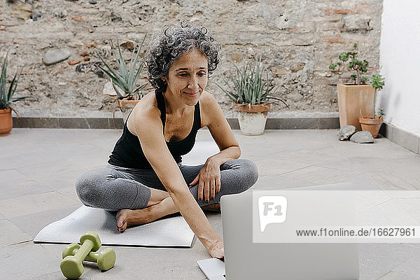 Lächelnde Frau mit Laptop für das Lernen Yoga durch Online-Tutorial  während im Hinterhof sitzen