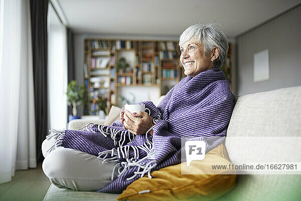 Lächelnde ältere Frau mit Decke  die eine Kaffeetasse hält  während sie zu Hause auf dem Sofa sitzt