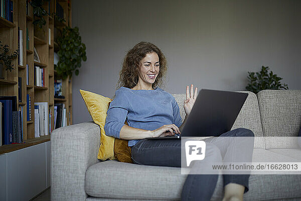 Mittlere erwachsene Frau winkt mit der Hand zum Videoanruf auf dem Laptop  während sie zu Hause sitzt