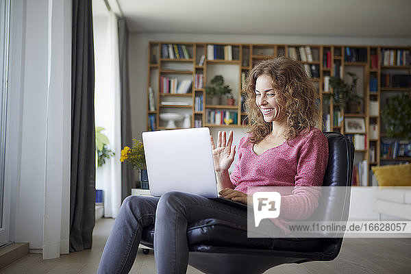 Lächelnde Frau winkt mit der Hand zum Videoanruf auf dem Laptop  während sie zu Hause sitzt