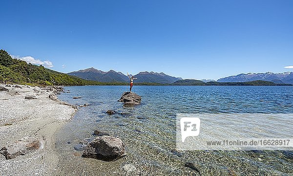 Junger Mann steht auf Stein im Wasser und streckt die Arme in die Luft  Strand Frasers Beach  Lake Manapouri  Manapouri  Südinsel  Neuseeland  Ozeanien