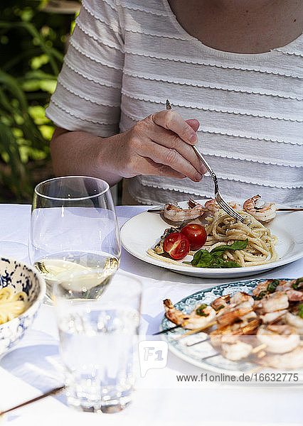Woman twirling pasta cacio e pepe on a plate with shrimp skewers  tomatoes and white wine at an outdoor table