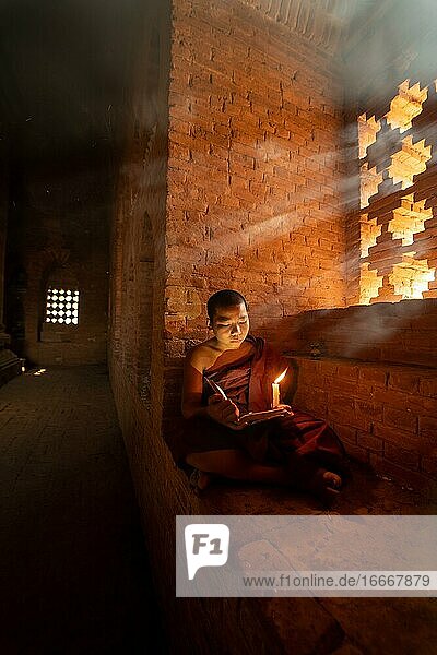 Buddhist young monk in red robe reading sitting in front of rays of light in a temple Bagan Myanmar Asia