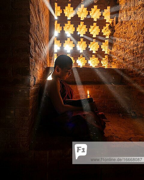 Buddhist young monk in red robe reading while sitting with a candle in front of rays of light in a temple Bagan Myanmar Asia