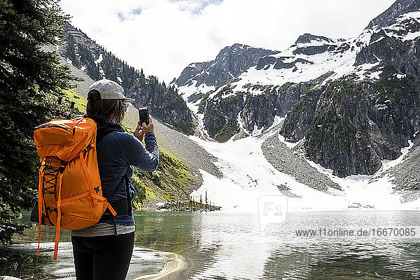 Eine Wanderin mit Rucksack und Smartphone steht in der Nähe eines Sees und fotografiert die malerische Landschaft der schneebedeckten felsigen Berge während einer Sommerreise in British Columbia