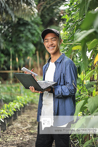 Junger asiatischer Bauer mit Laptop im Garten
