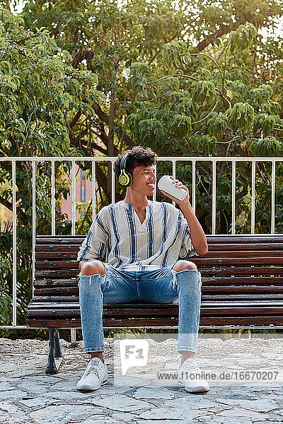 Young boy with afro hair drinks coffee while he is sitting on a bench